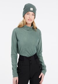 Pull à col roulé côtelé vert avec des manches longues associé à un pantalon noir taille haute avec des détails de boutons et des poches zippées.