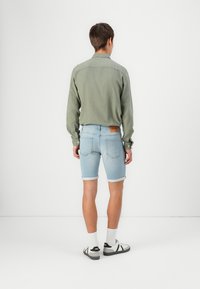 Camisa de manga larga verde claro, pantalones cortos de mezclilla en azul claro, con el dobladillo vuelto. Se lleva con calcetines blancos y zapatos con detalles negros. Vista trasera.
