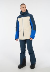 Farbig abgesetzte Skijacke in Beige, Marineblau und Blau, aus wasserdichtem Material mit Kapuze und Reißverschlusstaschen. Kombiniert mit marineblauen Hosen.