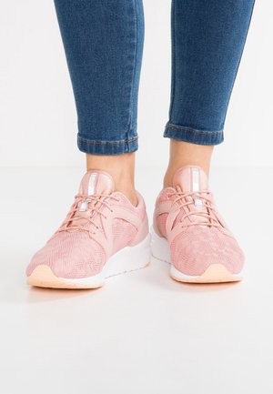Voeten met roze mesh sneakers met witte zolen en veters, gecombineerd met opgerolde blauwe spijkerbroek op een witte vloer.