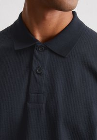 Navy textured polo shirt with a collar, three black buttons, and a subtle pattern. Features a side placket and smooth fabric.