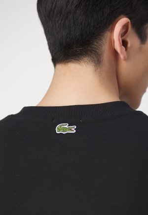 Maglione nero a maglia con scollatura a costine, caratterizzato da un piccolo logo di coccodrillo verde ricamato sul retro. Texture liscia e design aderente.