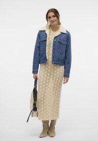 Veste en denim avec un col en fausse fourrure, associée à une robe en crochet beige. La tenue est complétée par des bottes beiges jusqu'aux genoux et un sac à bandoulière noir.