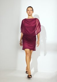 Diep bordeauxrode jurk met korte mouwen en een gerimpelde taille, gemaakt van lichte stof, gecombineerd met zwarte hakken sandalen.