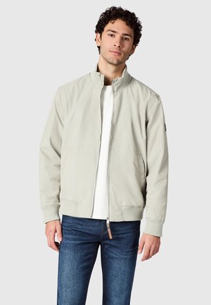 Junger Mann, der eine hellbeige Jacke mit Reißverschluss über einem weißen Hemd und blauen Jeans trägt, vor einem schlichten hellgrauen Hintergrund stehend.