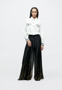 Biała koszula na guziki z kieszeniami i dodatkami na rękawach w paski zebry, zestawiona z czarnymi plisowanymi spodniami o szerokich nogawkach, posiadającymi subtelny połysk i wzorzysty materiał.