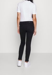 Persona de pie de espaldas, vistiendo una camiseta blanca de manga corta, jeans ajustados negros y zapatillas blancas sobre un fondo sencillo.