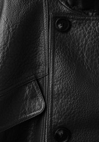 Black leather jacket close-up showing textured material, two buttons, and a flap pocket with detailed stitching.