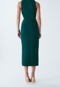 Vrouw in een mouwloze donkergroene midi-jurk en gouden hogere hakken, staand met haar rug naar de camera.