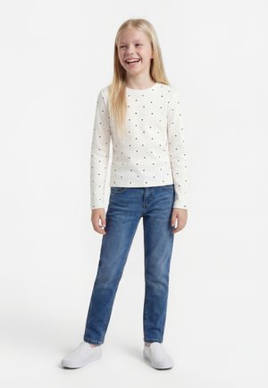 Fată stând și zâmbind, purtând o bluză albă cu mâneci lungi cu inimioare negre mici, blugi albaștri și pantofi slip-on albi.