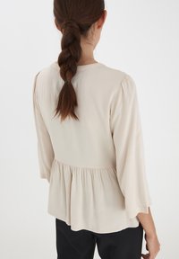 Blusa beige chiaro con maniche a tre quarti, scollo rotondo e vita arricciata. Tessuto morbido con una caduta fluida.