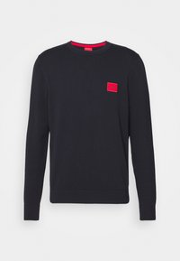 Granatowy sweter z okrągłym dekoltem wykonany z bawełny, z czerwonym naszywką na lewej piersi, ściągaczami przy rękawach i dolnej krawędzi. Gładka tekstura.