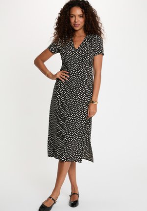 Mujer de pie con la mano en la cadera, vestida con un vestido midi negro con lunares blancos, mangas cortas y zapatos Mary Jane negros.