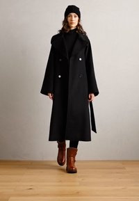Schwarzwollmantel mit einem breiten Kragen und doppelter Knopfleiste, kombiniert mit einem schwarzen Rollkragenpullover, schwarzen Leggings und braunen Stiefeln.