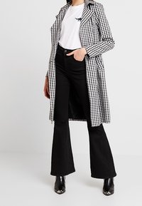 Musta-valkoinen ruudullinen trench-takki, jossa on leveät kaulukset, päällä valkoinen T-paita ja leveät mustat farkut. Mustat korolliset nilkkurit.