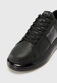 Sneaker in pelle nera con punta rotonda, chiusura con lacci, superficie testurizzata e suola lucida nera. Presenta il nome del marchio impresso sulla linguetta e sul lato.