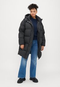 Black puffer coat with a hood, quilted design, and snap button accents. Worn over a navy sweater and blue flared jeans, featuring black chunky boots.