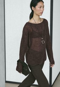 Bruine doorschijnende gebreide trui met brede mouwen, gecombineerd met donkerbruine broek. Accessoires zijn onder meer een donkergroene, getextureerde handtas en een zilveren ketting.