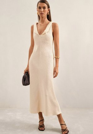 Cremfarbendes ärmelloses Kleid mit einem strukturierten Muster, tiefem V-Ausschnitt und fließender Silhouette, kombiniert mit einer schwarzen Handtasche und Riemchenschuhen.