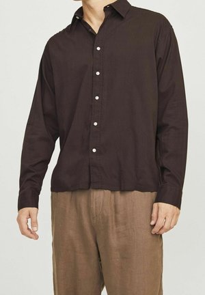 Hombre vestido con una camisa de manga larga marrón oscuro abotonada y pantalones sueltos marrón claro, de pie frente a un fondo claro y liso.