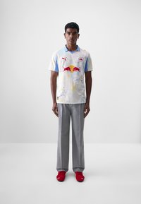 Puma sportska majica u svijetlim pastel bojama, s logotipom Red Bulla i cvjetnim uzorcima, uparena sa sivim hlačama i crvenim cipelama.