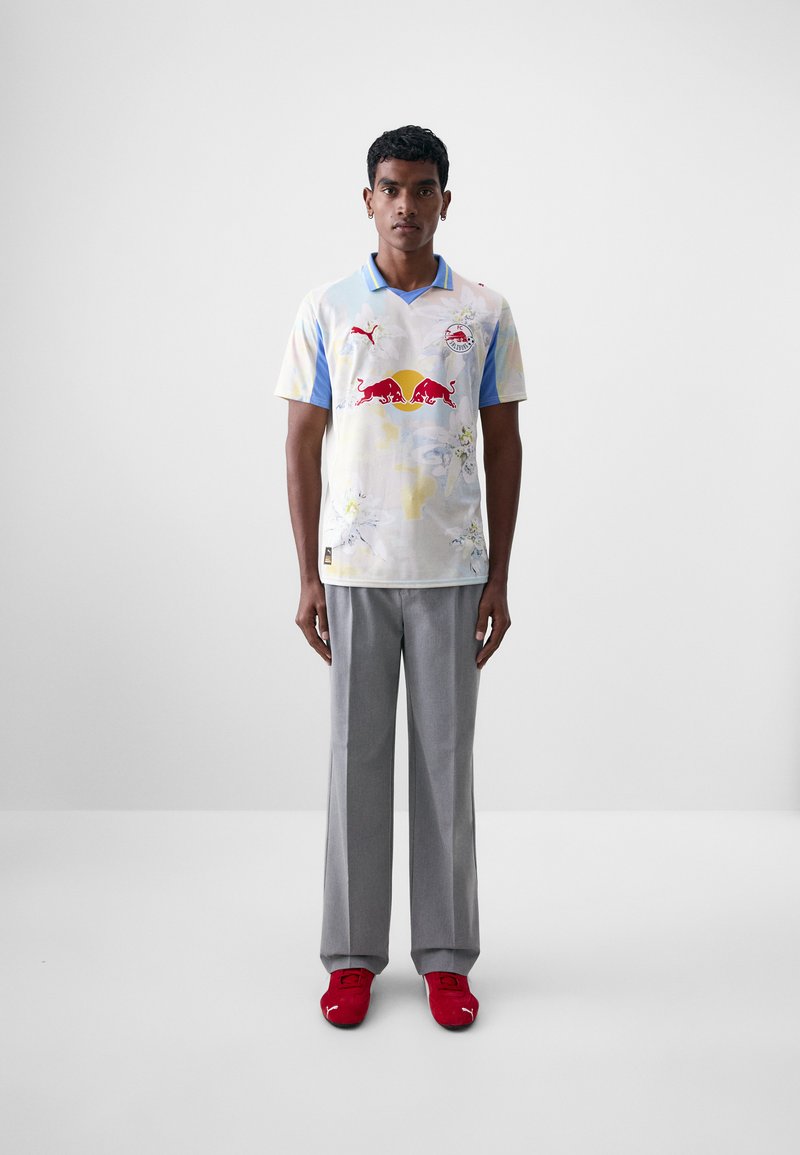 Puma sportska majica u svijetlim pastel bojama, s logotipom Red Bulla i cvjetnim uzorcima, uparena sa sivim hlačama i crvenim cipelama.