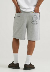 Pantaloni corti in jersey grigi con tasche laterali. Presentano una grafica di un scheletro cartoon sulla tasca posteriore. Orli rifiniti e vestibilità rilassata. Accenti in metallo neri.