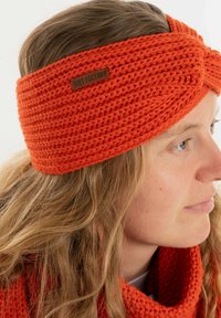 Gehaakte oranje hoofdband met een gedraaide ontwerp en dikke, gestructureerde garen. Een klein bruin leren label is aan de zijkant bevestigd.