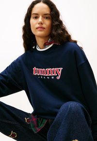 Φούτερ σε ναυτικό μπλε με την επιγραφή "tommy" κόκκινα και "JEANS" άσπρα, με στρογγυλή λαιμόκοψη και ριμπ υφή, φορεμένο πάνω από ένα πουκάμισο καρό.