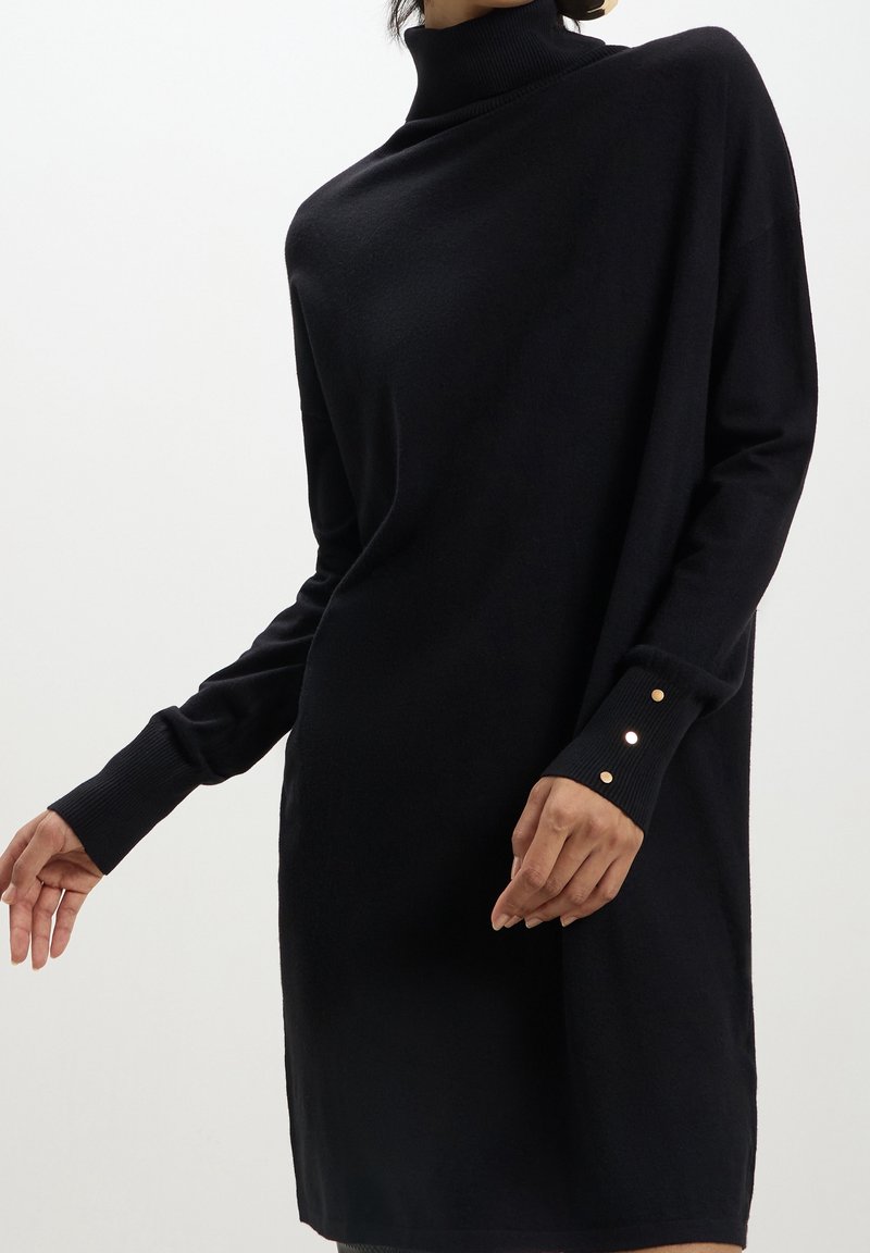 Femme portant une robe pull à col roulé noire à manches longues avec des boutons dorés sur le poignet, sur un fond blanc.