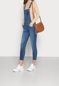 Jeanslatzhose mit dunkelblauer Waschung, tailliertem Schnitt und Taschen; kombiniert mit einem beigen Rippenoberteil und weißen Sneakers; braune Umhängetasche.