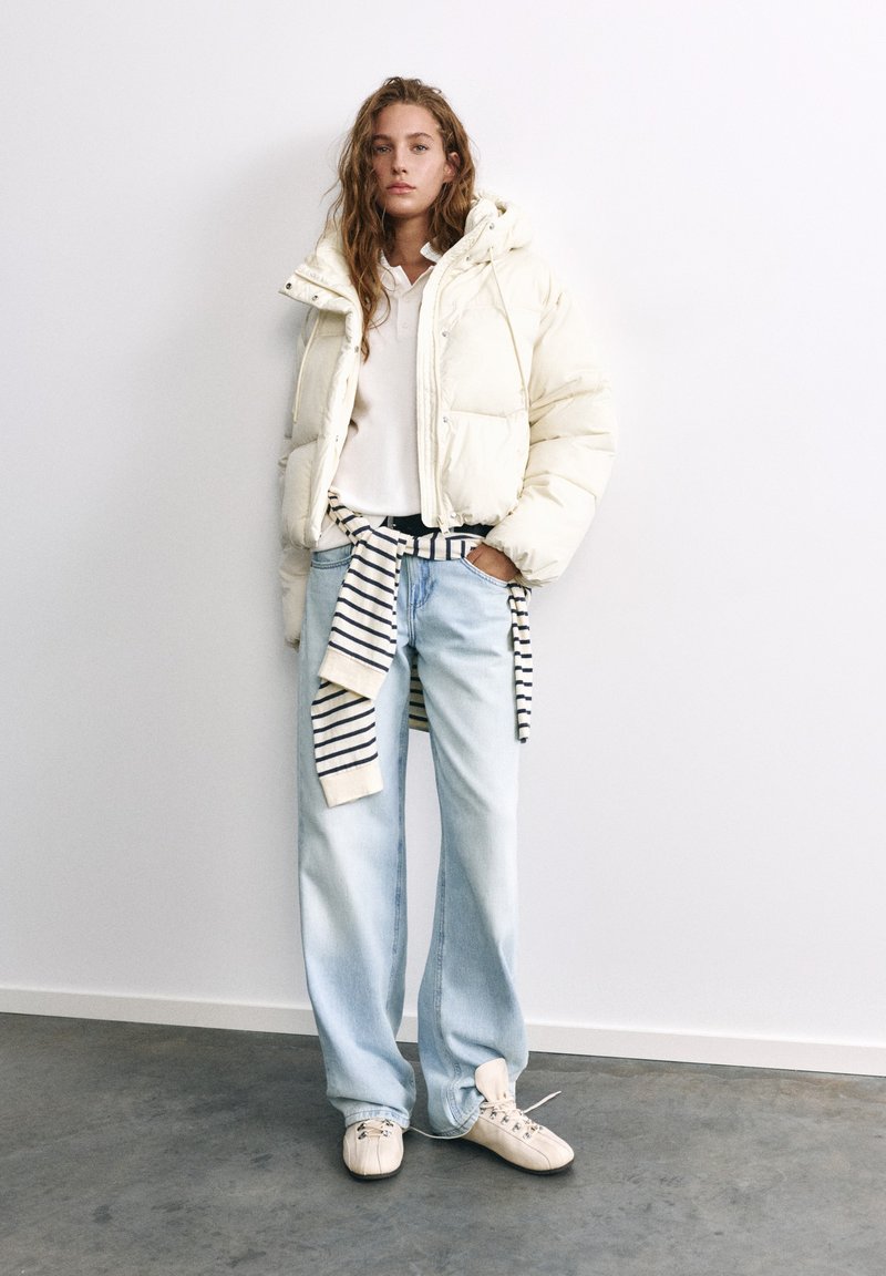 Jeune femme portant une doudoune blanche, un jean large bleu clair, des baskets blanches et un pull rayé noué autour de la taille.