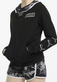 Sudadera negra con cremallera, con capucha forrada en camuflaje gris, bolsillos frontales y logotipo de Puma en blanco en el pecho y las mangas. Combinada con pantalones cortos de camuflaje.