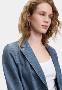 Giacca in denim di un blu chiaro, con revers a scollatura a V, vestibilità rilassata e texture morbida. Indossata sopra una canotta bianca. Accentuata da cuciture pulite.