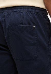 Κοντινό πλάνο ενός ατόμου που φοράει μπλε σκούρο παντελόνι με ελαστική μέση και πίσω τσέπη με μικρή ασπρόμαυρη ετικέτα.