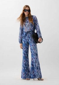 Blue patterned outfit featuring long-sleeve top and flared pants in a smooth fabric, paired with a black handbag and oversized sunglasses.
