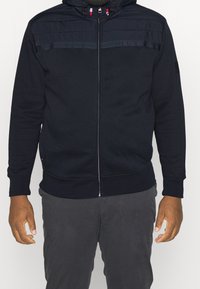 Navy zip-up hoodie with a textured shoulder panel, adjustable drawstring hood, and ribbed cuffs and hem, made from soft cotton blend fabric.