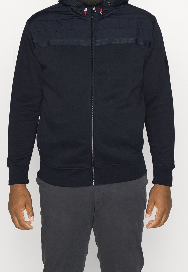 Navy zip-up hoodie with a textured shoulder panel, adjustable drawstring hood, and ribbed cuffs and hem, made from soft cotton blend fabric.