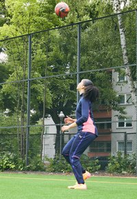 Sportaš u tamnoplavom i roza sportskom odijelu skače kako bi uhvatio narančastu i crnu nogometnu loptu na zelenom terenu, okružen žičanom ogradom.