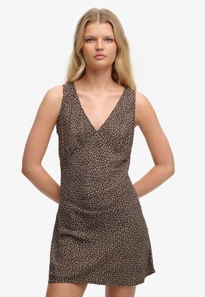 Ung kvinde med langt blondt hår iført en brun og beige ærmeløs kjole med blomsterprint og V-hals, stående med hænderne bag ryggen.