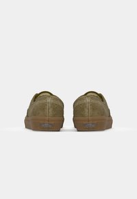 Olivengroene suède schoenen met witte stiksels en een rubberen zool; achteraanzicht. Kenmerkt het Vans-logo op de hak en een ronde vorm.