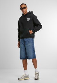Zwarte hoodie met voorzak, voorzien van een witte scriptlogo. Gecombineerd met denim shorts en grijze sportschoenen.