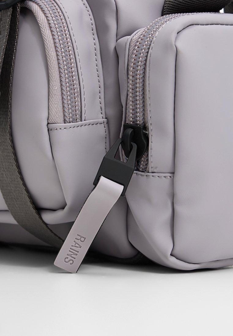 Lichtgrijze rugzak van waterdicht materiaal, met dubbele ritsen, een lusvormige band met "RAINS"-tekst en getextureerde accenten.
