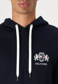 Tamno plava dukserica s bijelim vezicama, s izvezenim grbom s lavovima i tekstom "HILFIGER" na prsima. Mekani materijal.