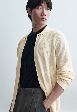 Fiatal személy rövid fekete hajjal, bézs mintás kardigánt visel fekete bordázott galléros póló fölött és szürke nadrágot, oldalra nézve.