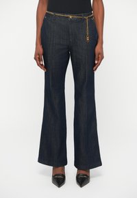 Un pantalon en denim foncé évasé avec une taille haute, une accentuation en chaîne dorée et des surpiqûres contrastantes, associé à des chaussures noires pointues.