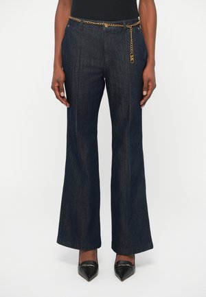 Un pantalon en denim foncé évasé avec une taille haute, une accentuation en chaîne dorée et des surpiqûres contrastantes, associé à des chaussures noires pointues.