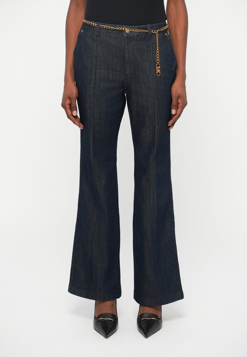Un pantalon en denim foncé évasé avec une taille haute, une accentuation en chaîne dorée et des surpiqûres contrastantes, associé à des chaussures noires pointues.