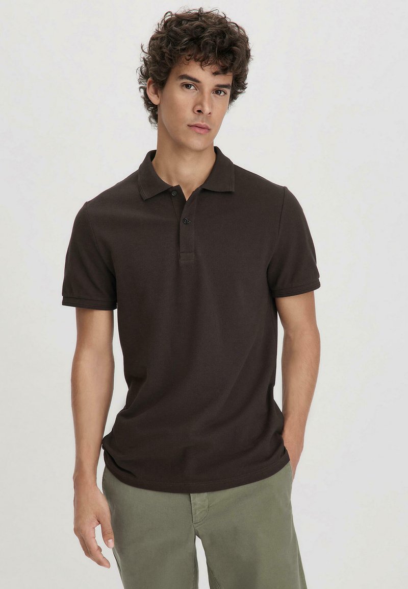 Brown polo shirt made of cotton, featuring a collar and three-button placket. Slim fit with short sleeves, paired with light green pants.
