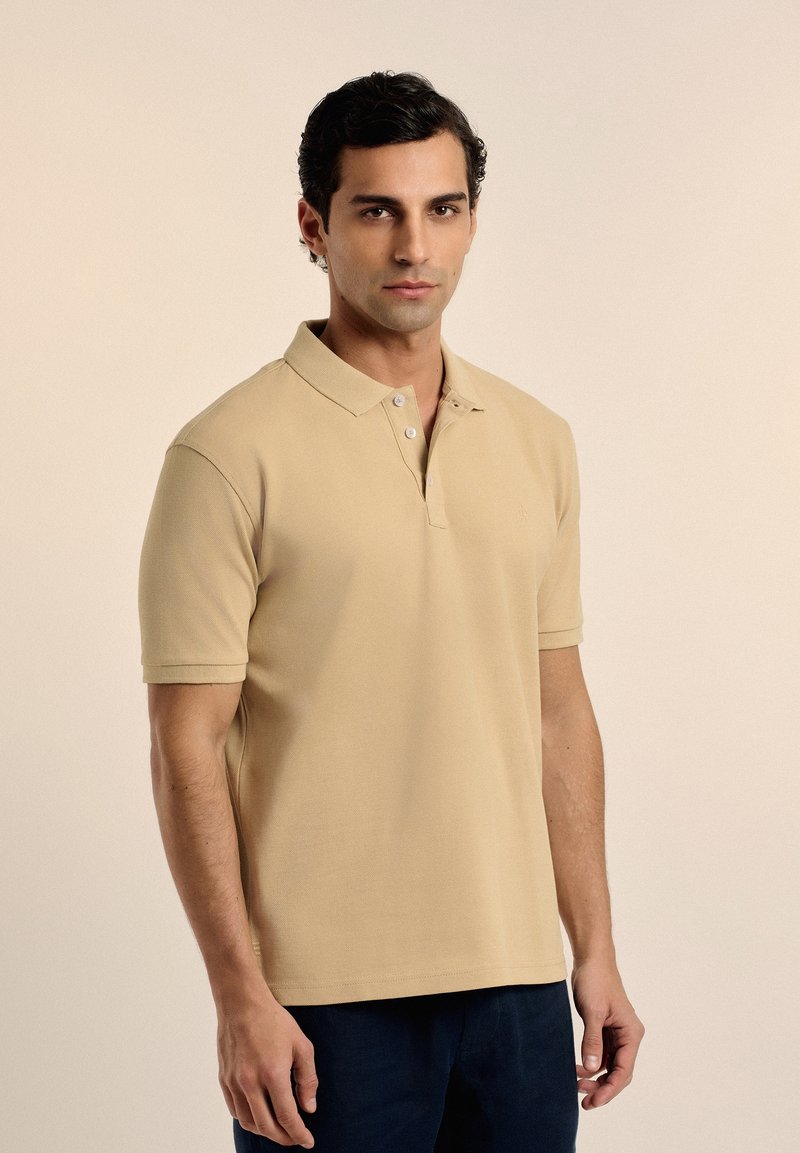 Camisa polo beige hecha de tejido texturizado, con una tapeta de dos botones, mangas cortas y un sutil logo en el pecho.